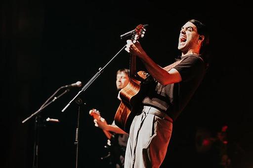 A musician passionately sings and plays guitar on stage, while another musician stands behind him.