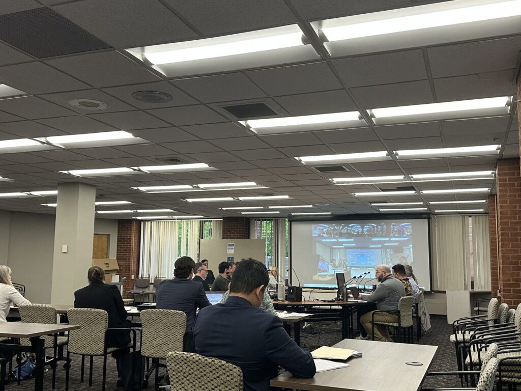 A group of people seated at tables, discussing in a meeting room with a video screen displaying participants.
