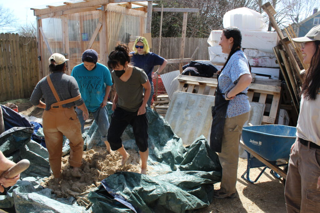 A group of people mix materials in a large tarp, with a wooden structure and tools in the background.