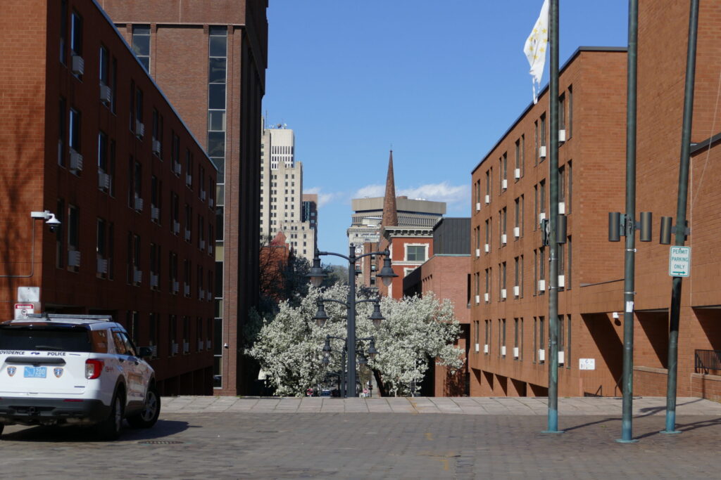 A police vehicle is parked on a street lined with brick buildings and blooming trees, with city buildings in the background.