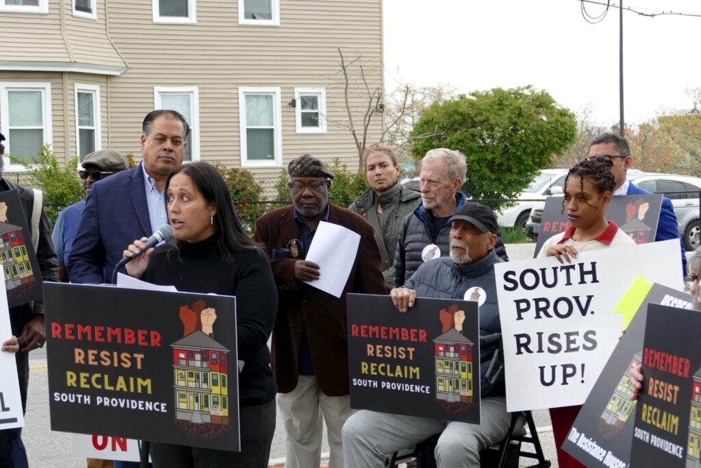 A speaker addresses a crowd holding signs advocating for South Providence.