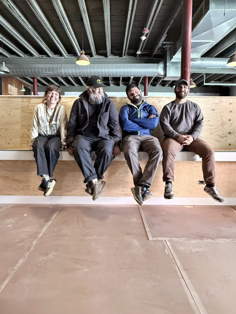 Four people sit on a ledge, smiling and relaxed, in a spacious interior with exposed beams and lights.