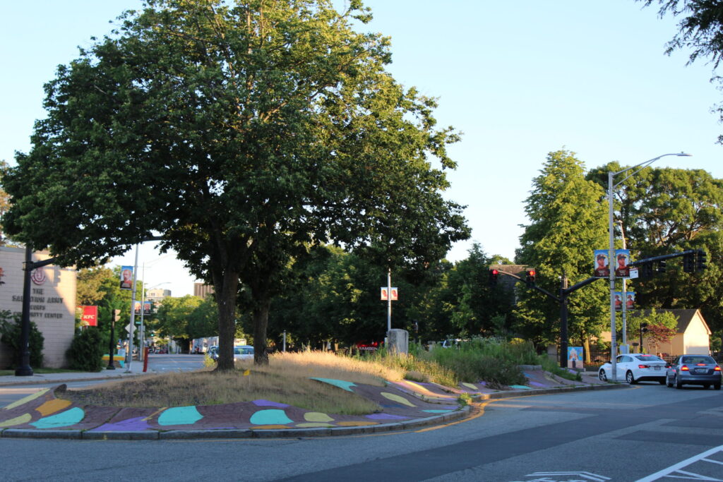 A large tree stands beside a colorful traffic island, with cars and traffic lights in the background.