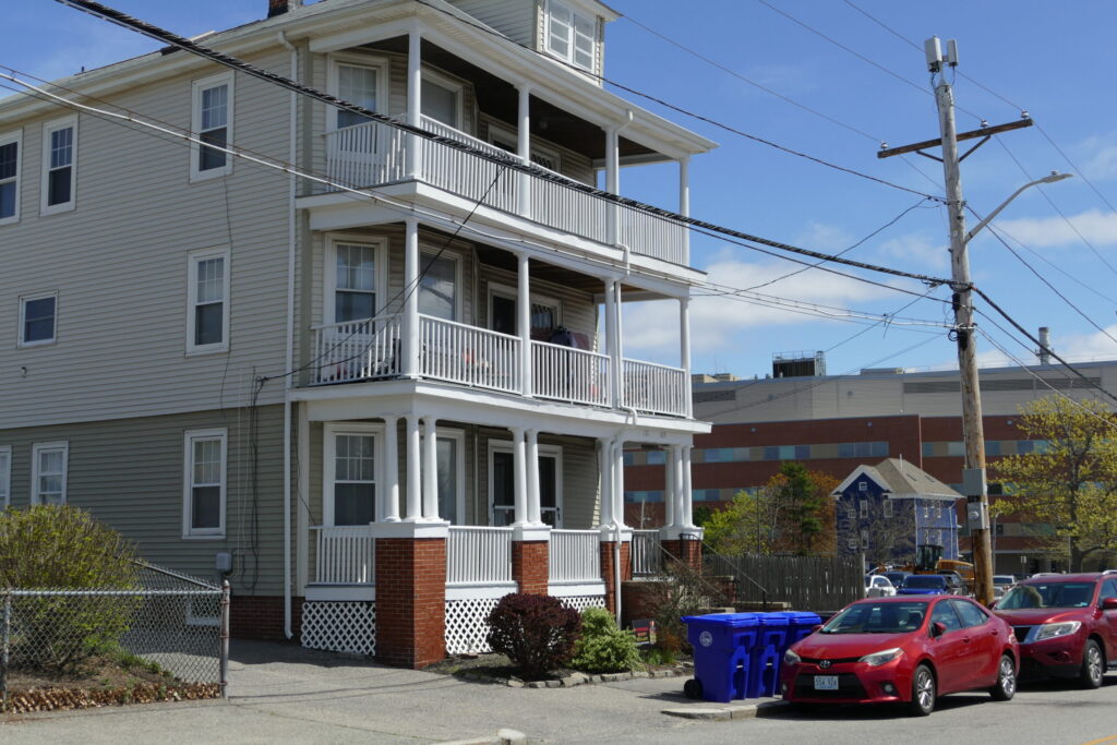 Three-story residential building with balconies, parked red cars, and blue recycling bins in front.