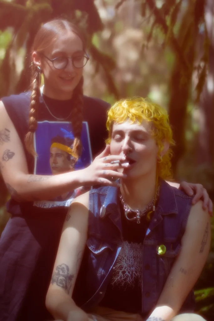Two people interact outdoors; one smiles while touching the other's face, who holds a cigarette. Both have visible tattoos.
