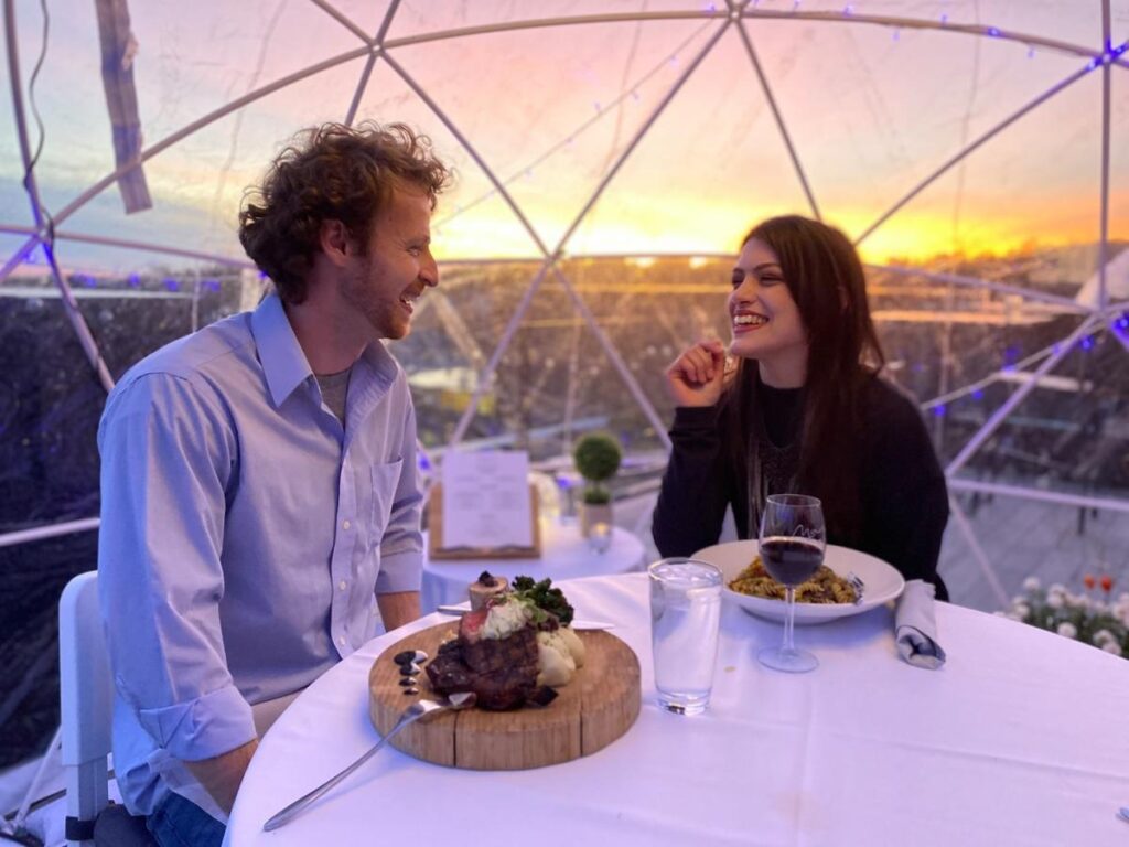 A man and woman share a meal in a cozy dome, smiling at each other with a sunset backdrop.