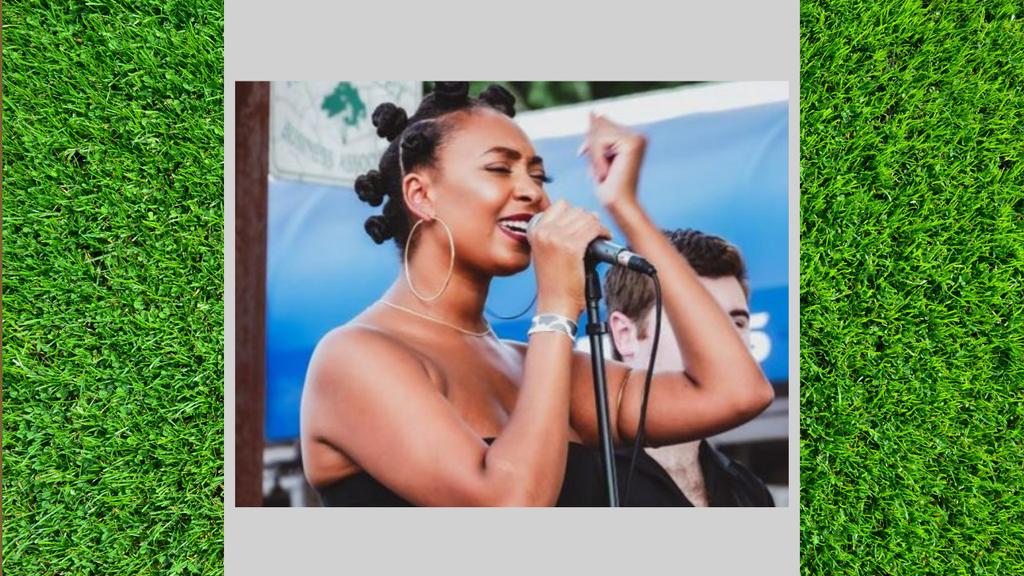 A woman sings passionately into a microphone, with a musician partially visible behind her.