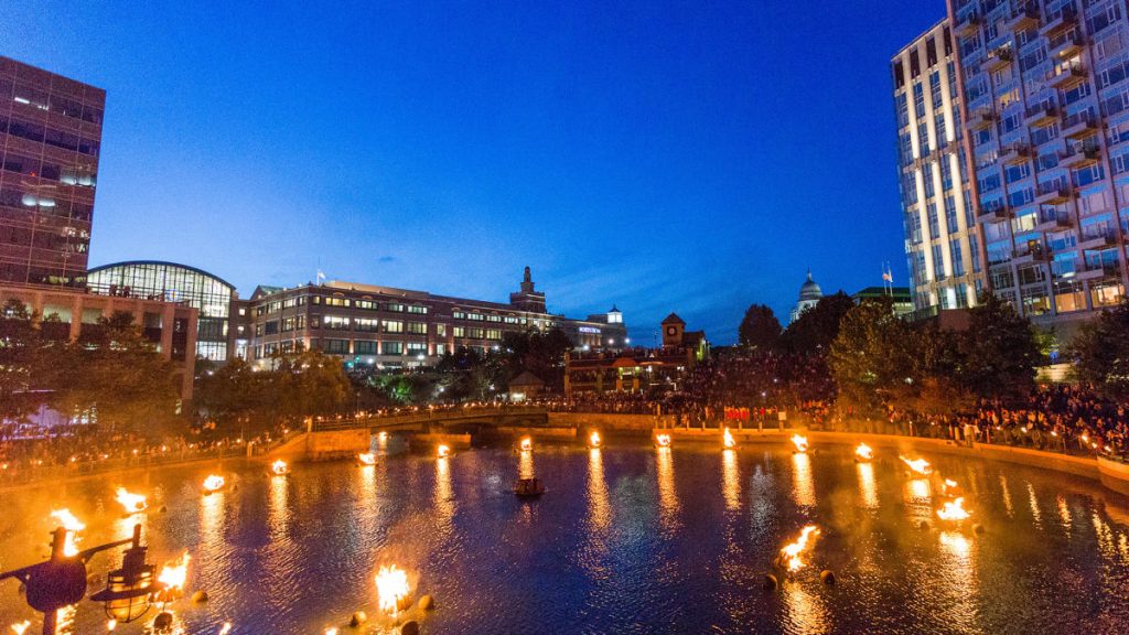 A waterfront scene at dusk with lit torches, buildings, and a large crowd gathered along the shore.