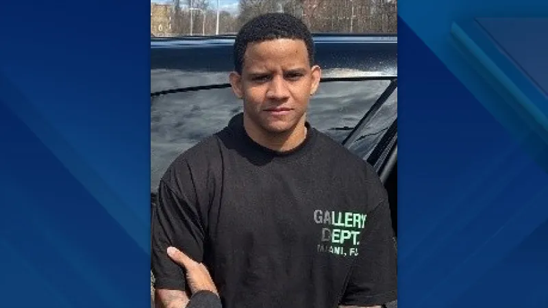 A man in a black t-shirt stands near a vehicle, looking directly at the camera.