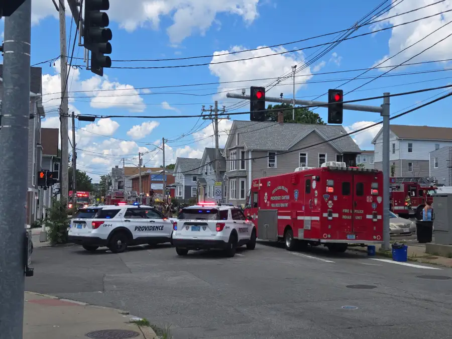 Emergency vehicles block the street at a fire scene, with police and fire units present under a blue sky.