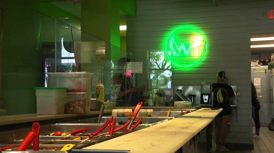 Brightly lit restaurant interior with a green logo, food prep area, and staff preparing meals in the background.