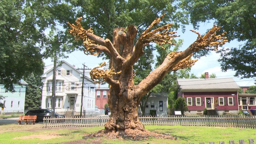 Large, artistic tree sculpture with intricate branches in a park, surrounded by houses and greenery.