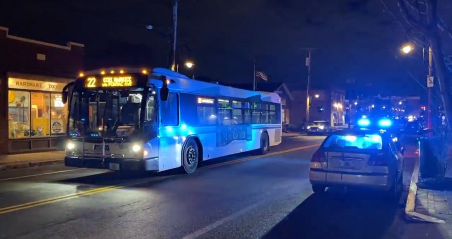 A city bus with blue lights approaches a scene with police vehicles and illuminated streetlights at night.