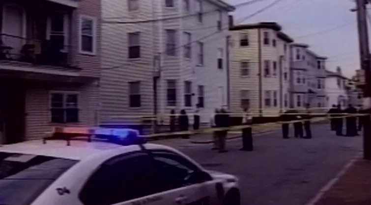 Police car with flashing lights parked near a crime scene surrounded by yellow tape and several officers.