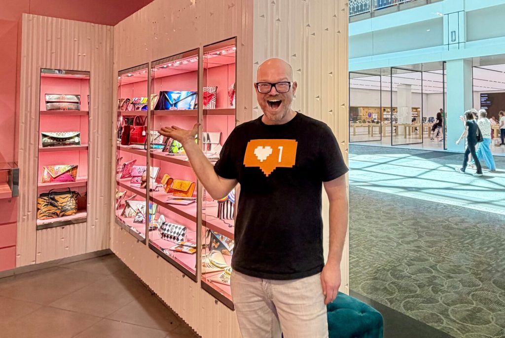 A smiling man in a black t-shirt gestures toward a display of colorful handbags in a pink-lit store.