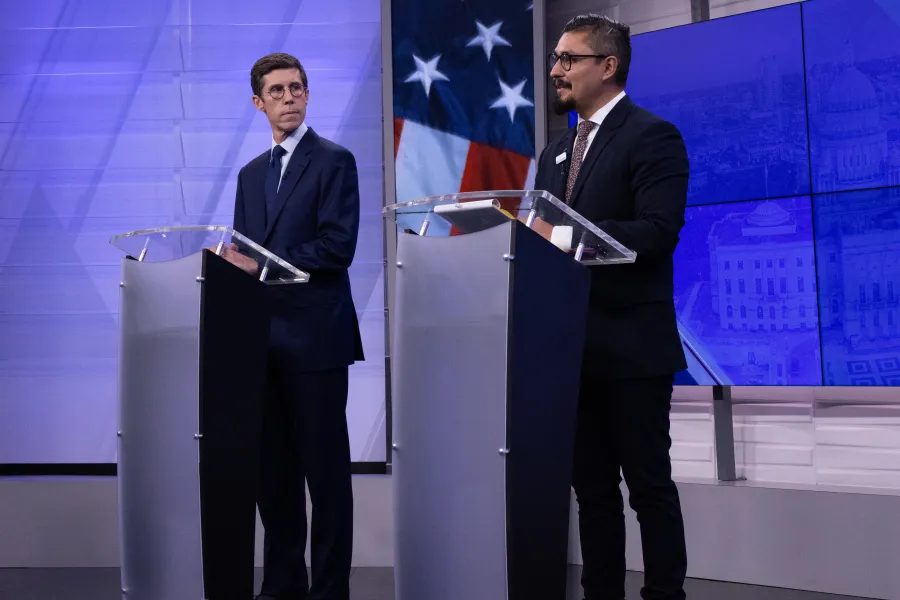 Two men stand at podiums during a debate, with a backdrop featuring an American flag.