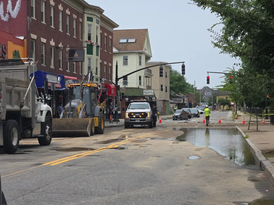 Construction vehicles and workers are present on a flooded street with traffic signals and nearby buildings.