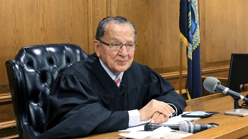 A judge sits at a wooden bench, wearing a black robe, with a microphone and papers in front of him.