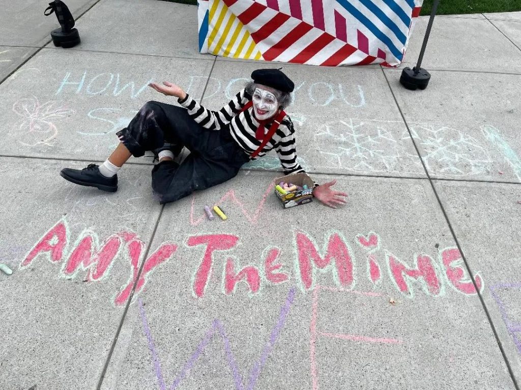 A mime in a striped shirt sits on pavement, surrounded by colorful chalk drawings, holding a box of chalk.