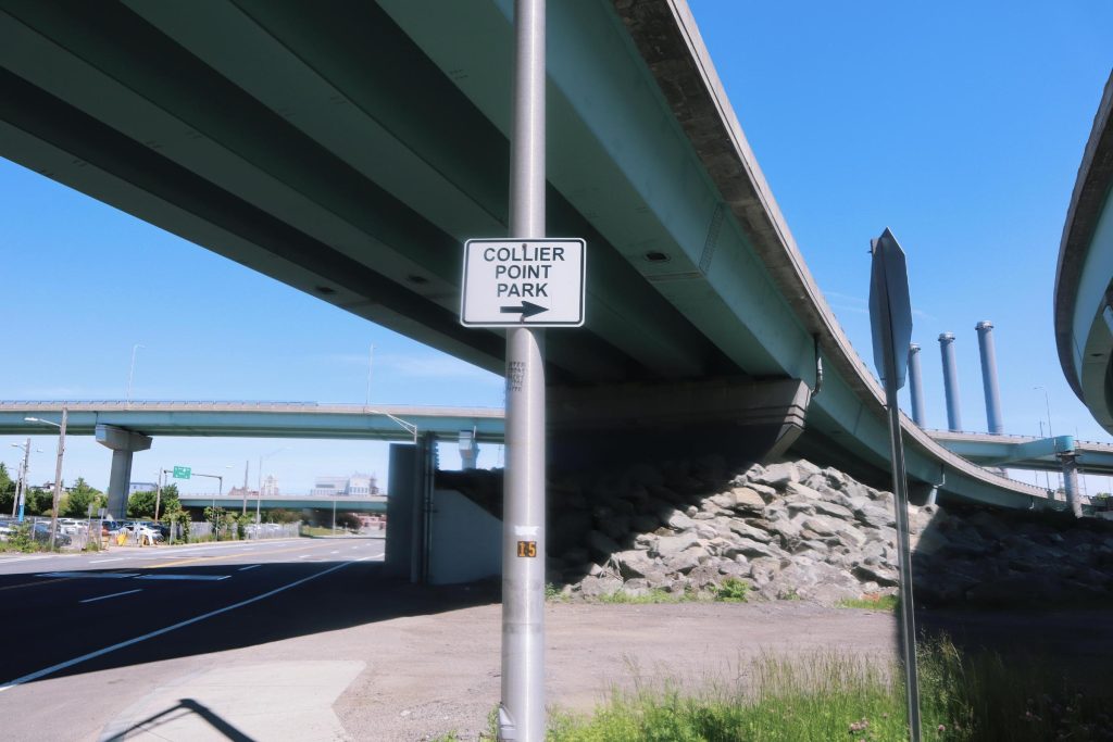 Sign directing to Collier Point Park under elevated highways, with rocky terrain and clear blue sky.