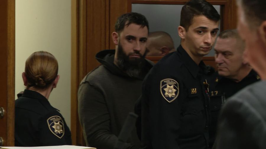 A man with a beard stands near police officers in a courtroom setting.