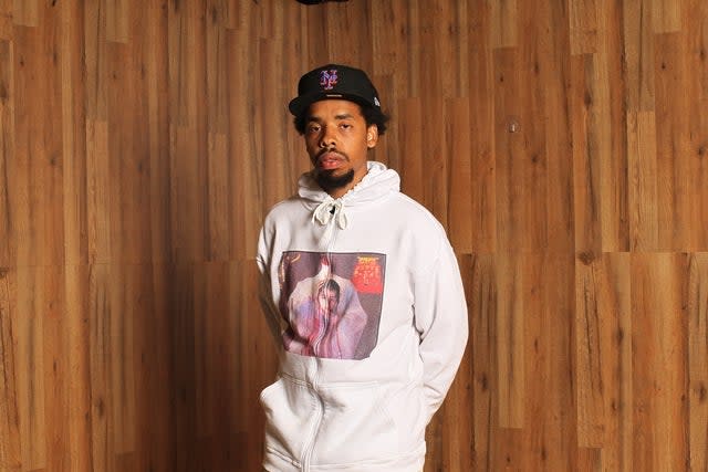 A man stands against a wooden backdrop, wearing a white hoodie with a graphic design and a black cap.