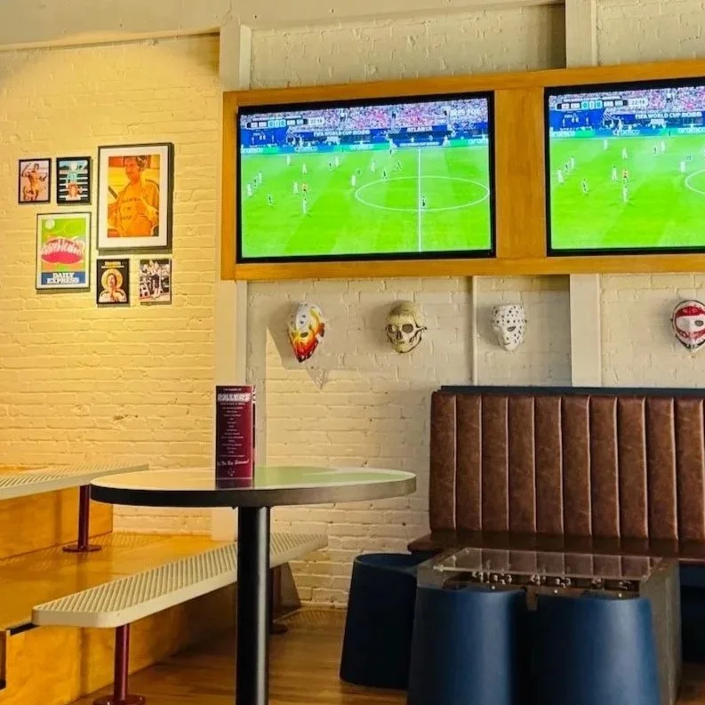 Two TVs display a soccer match. Decor includes framed art and masks. A table and seating area are visible.