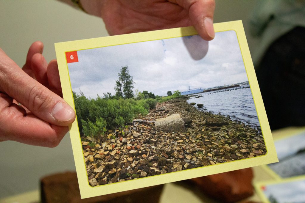 A person holds a photo of a rocky shoreline with vegetation and water in the background.