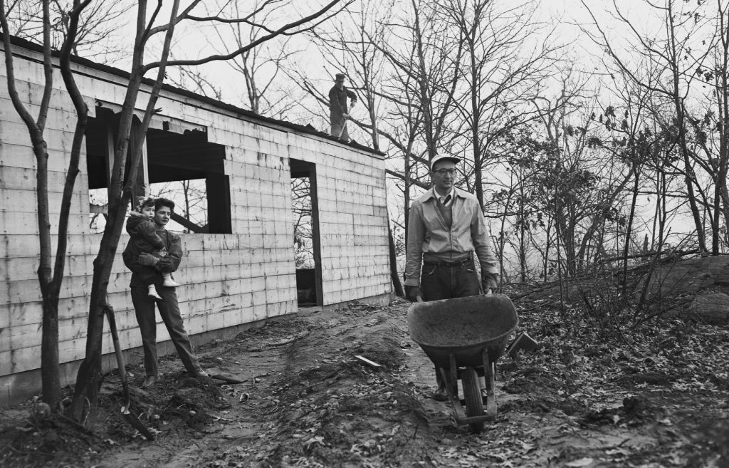 A man pushes a wheelbarrow while a woman holds a child near a partially constructed building. Another person works on the roof.
