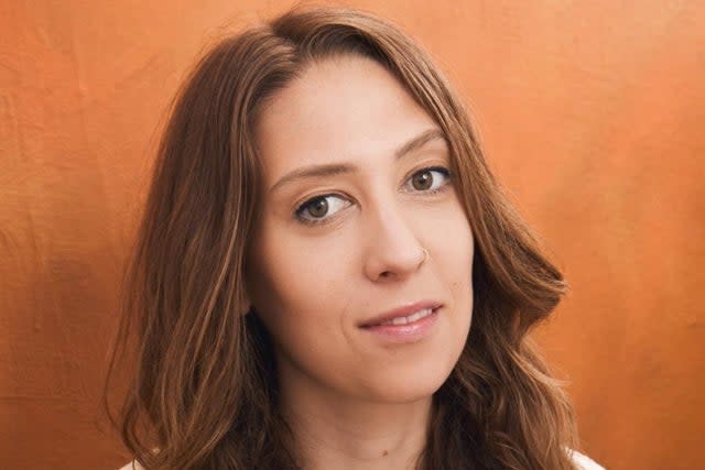 A woman with long, wavy hair poses against an orange background, looking directly at the camera.