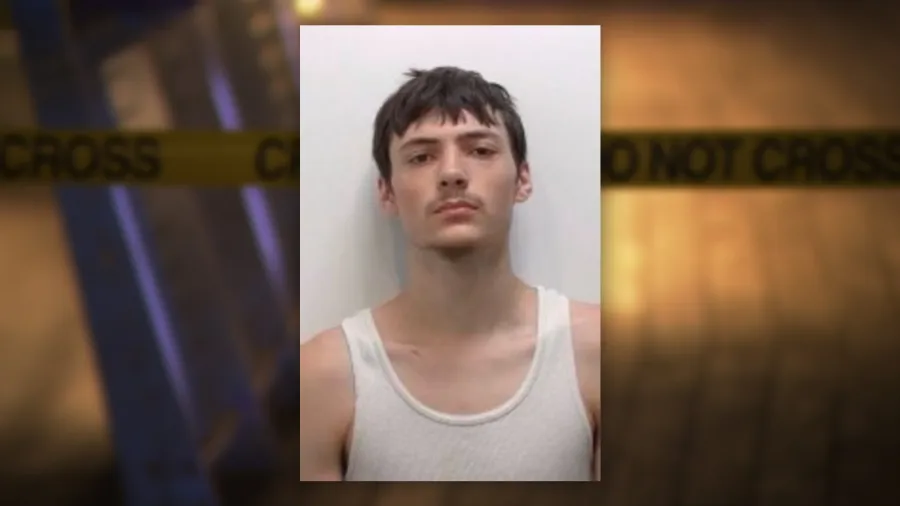 Booking photo of a young man with short hair and a mustache, wearing a white tank top. Background features police tape.