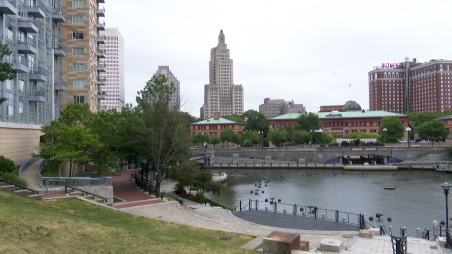 Cityscape featuring buildings, a river, and green trees under a cloudy sky. Pathways line the waterfront.