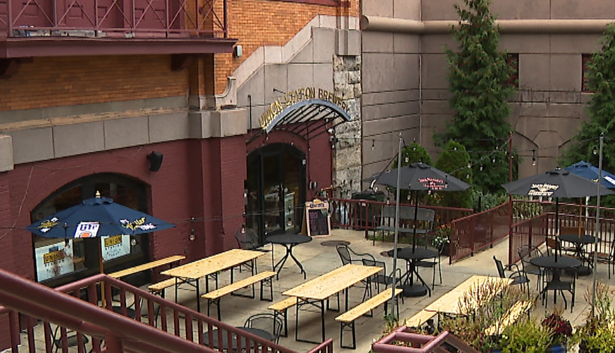 Outdoor seating area of a brewery with tables, umbrellas, and greenery. Entrance visible in the background.