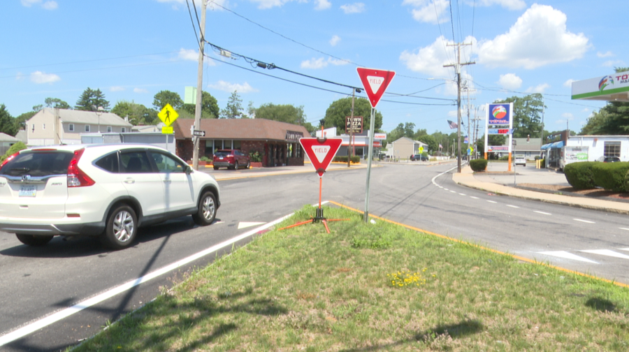 A white SUV approaches a yield sign at a road intersection with businesses nearby.