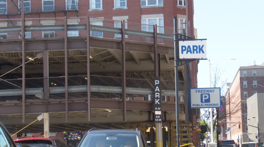 Parking garage with multiple signs indicating parking availability and rates. Nearby buildings visible in the background.