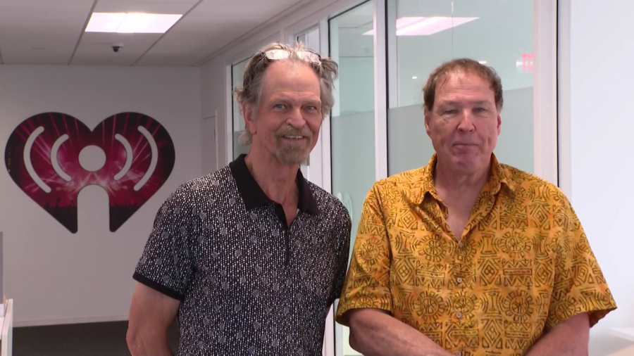 Two men stand side by side in a modern office, smiling. A large iHeartRadio logo is visible in the background.