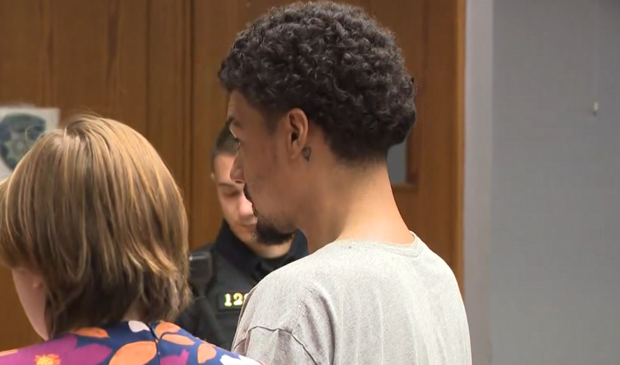 Two individuals stand in a courtroom, one with curly hair and a tattoo, the other with short hair and a floral shirt.