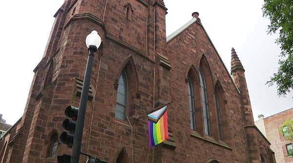 Historic stone church with a rainbow flag hanging from the side and traffic lights in the foreground.