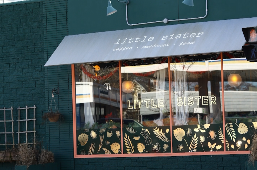 Exterior of "Little Sister" café, featuring large windows with decorative plants and a sign displaying the café's name.