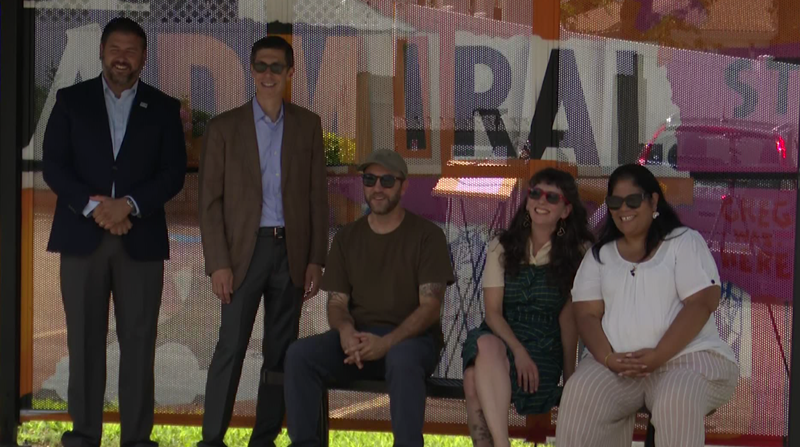 Five people pose together in front of a colorful mural, smiling and seated on a bench.