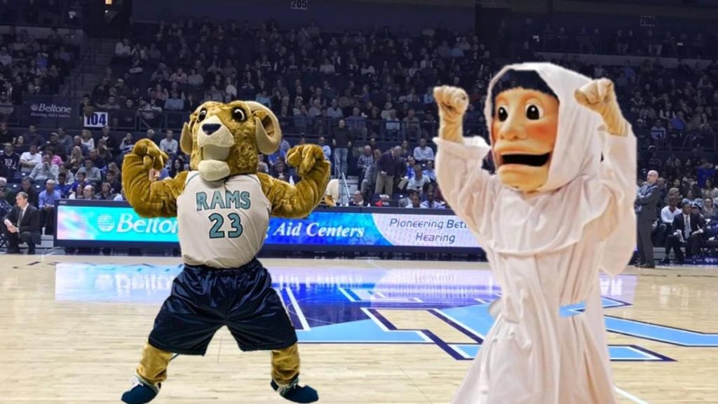 Two mascots celebrate on the basketball court, one dressed as a ram and the other in a white robe. Fans watch in the background.