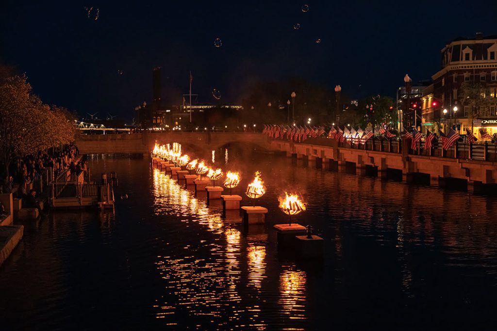 A river illuminated by floating braziers, with spectators along the banks and American flags in the background.