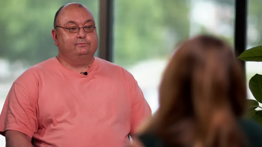 A man in a pink shirt sits facing a woman, engaged in conversation. Green foliage is visible in the background.
