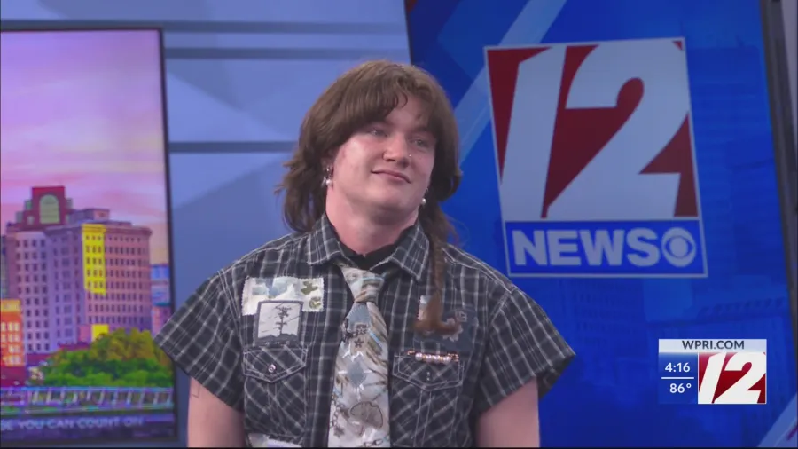 A person with long hair wears a patterned shirt and tie, standing in front of a news backdrop.