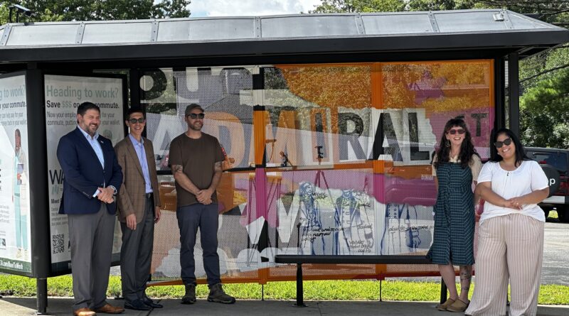 Five people stand at a bus stop, smiling. Colorful artwork decorates the shelter behind them.