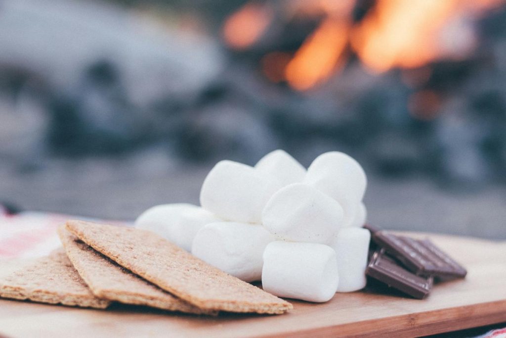 Graham crackers, marshmallows, and chocolate arranged on a wooden surface, with a fire in the background.