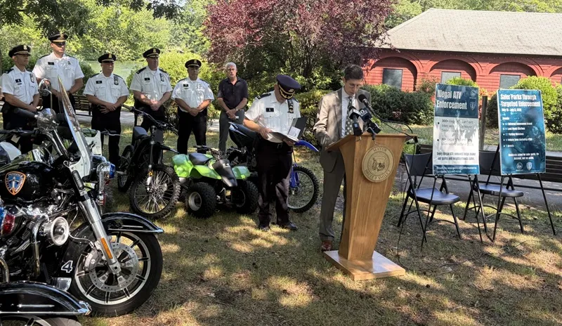 Officials gather at a podium for an ATV enforcement announcement, flanked by police and vehicles.