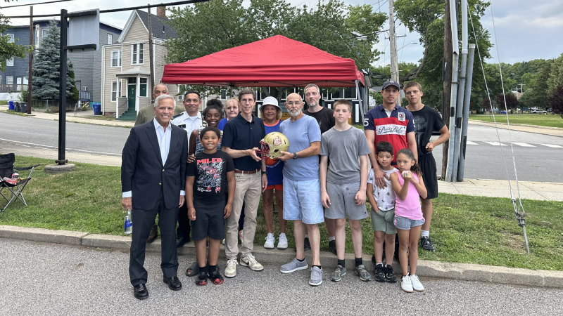 Community members gather under a tent, holding a basketball after replacing stolen youth helmets.