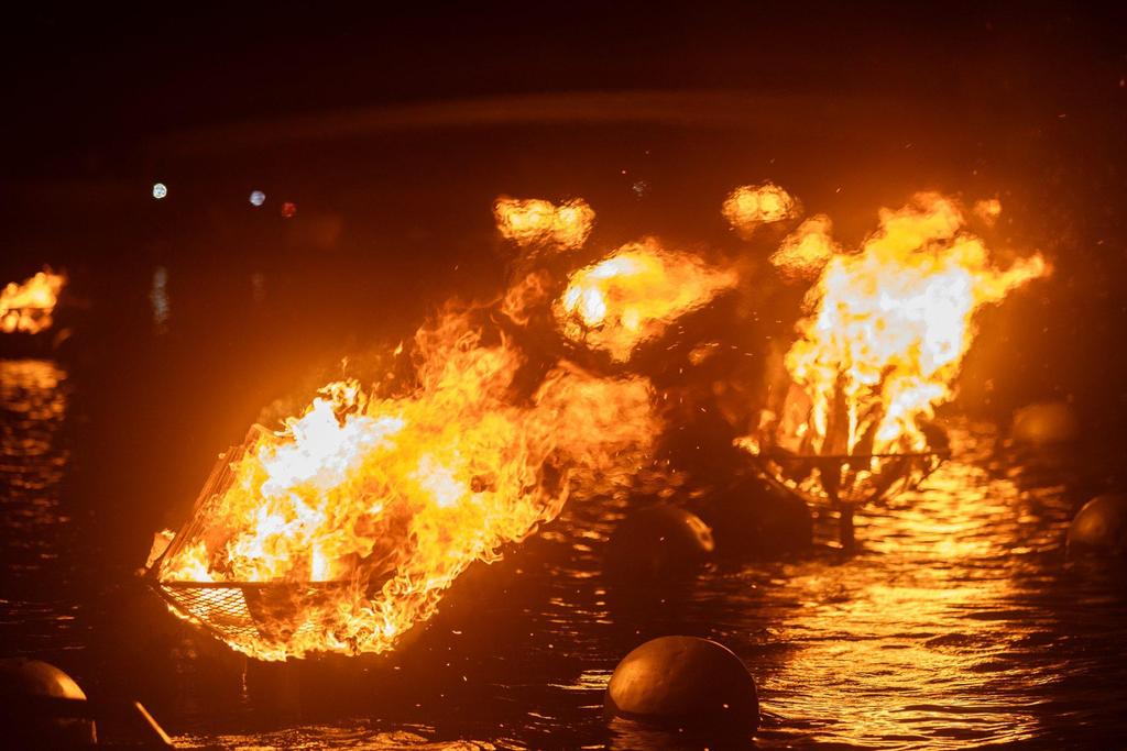Flames erupt from floating baskets on a dark water surface, illuminating the night.