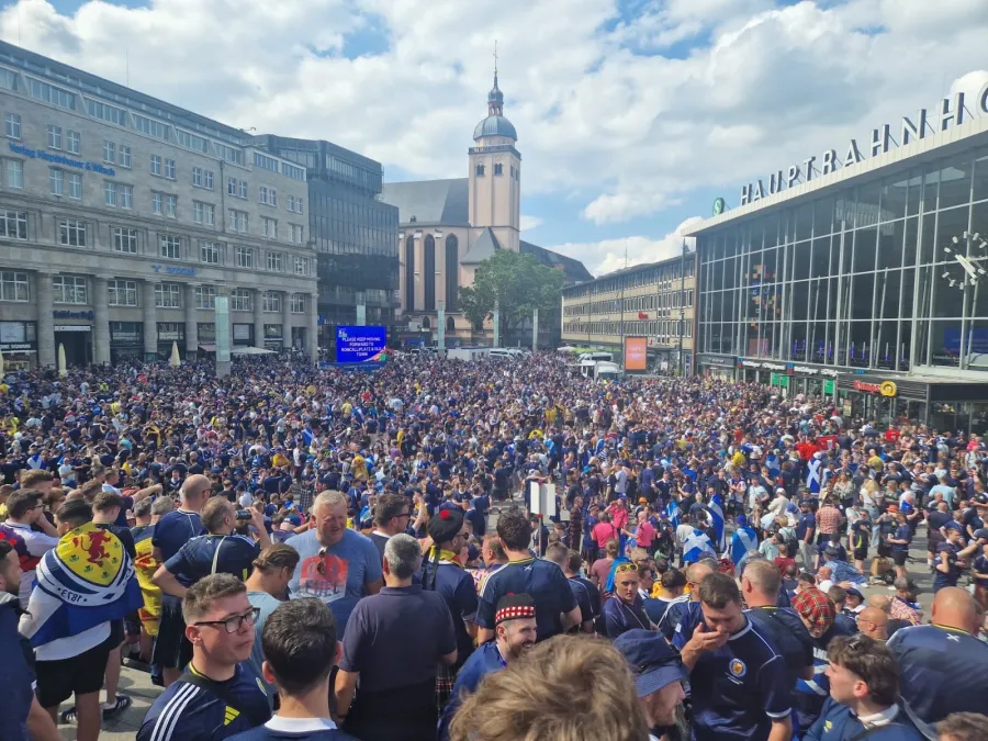 Crowd of fans in blue and yellow shirts gather in a city square, with buildings and a church in the background.